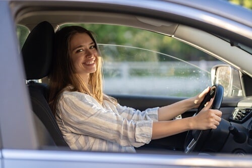 24 hour pick up and drop off services with Columbia Auto Care and Car Wash in Columbia MD. Image of young attractive woman smiling while sitting in driver side of car holding steering wheel on a sunny day with window lowered 3/4 of the way down.