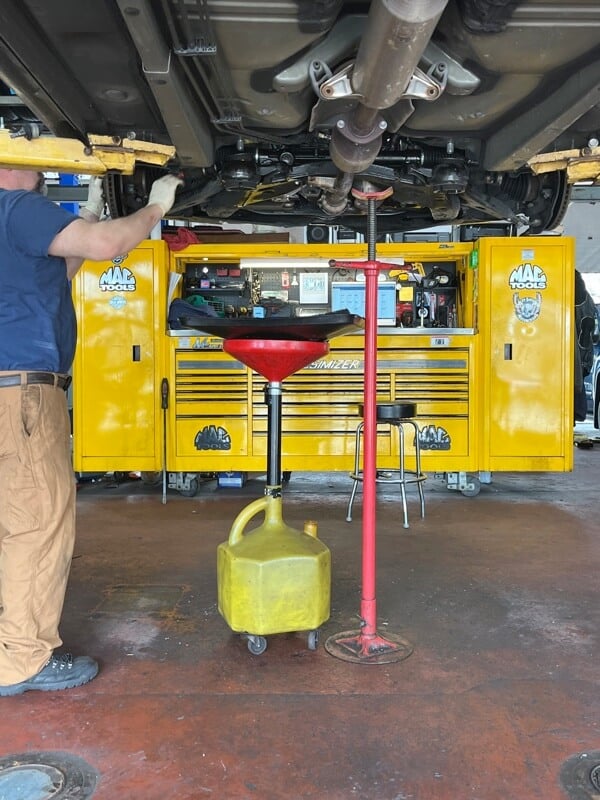 ASE certified mechanic at Columbia Auto Care in Columbia MD working underneath car getting oil change in columbia md shop with oil drain catching tool and large yellow tool box in background