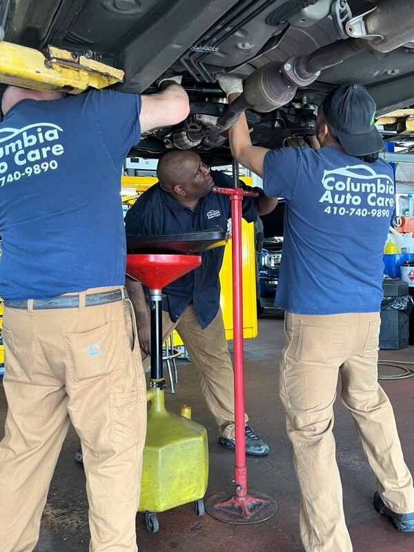 Three ASE certified mechanics that work at Columbia Auto Care in Columbia MD. working together under car that is on lift in shop bay