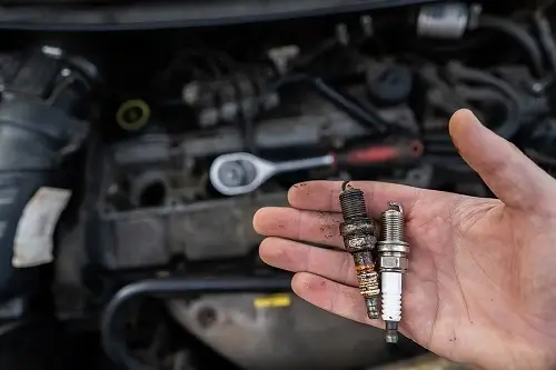 Does Your Car Need a Tune-Up? | Columbia Auto Care & Car Wash in Columbia, MD. Closeup image of old and new spark plugs in the hands of a mechanic with a car engine at the background. Concept image of car tune-up.