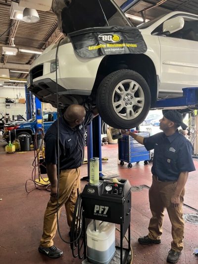 general car maintenance in columbia md being performed by 2 ASE certified mechanics at Columbia Auto Care; they are discussing issues they see standing underneath small white suv on lift in sop with diagnostic tool next to them as well as BG Services fender cover on car near engine with open hood